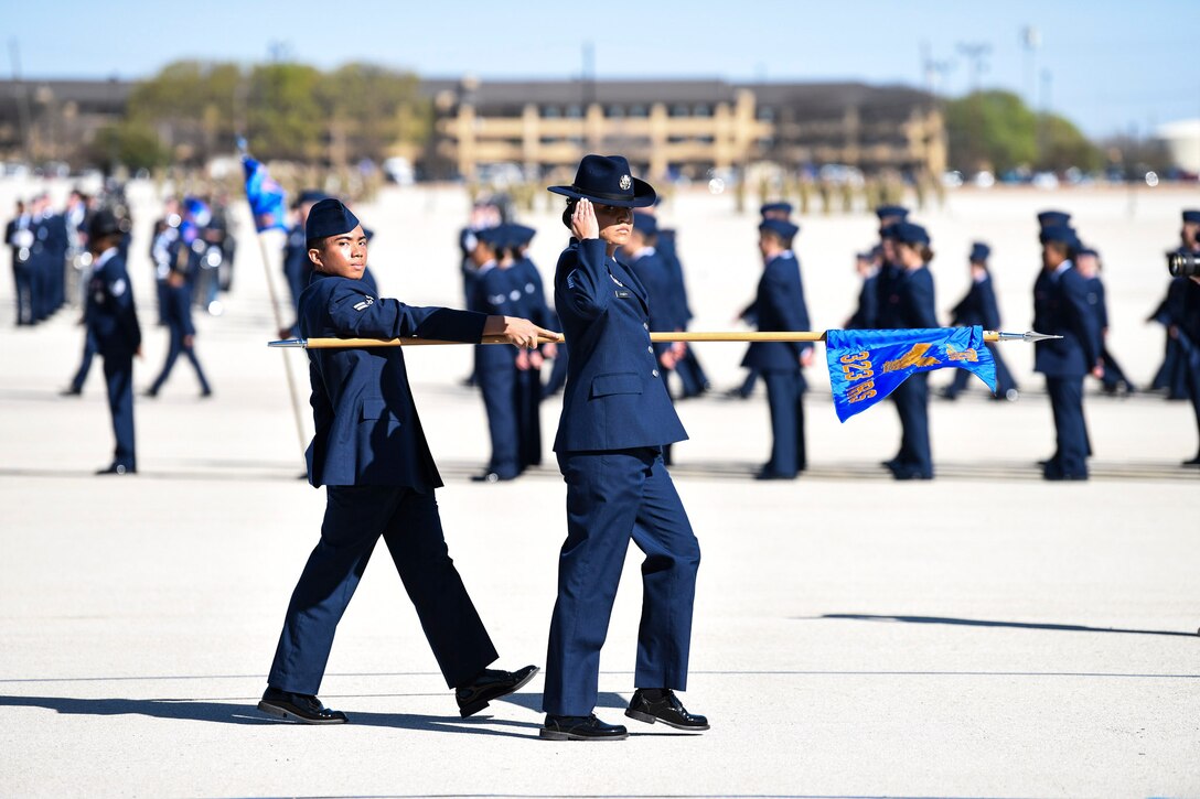 Two service members hold a flag and  walk in formation in front of others.