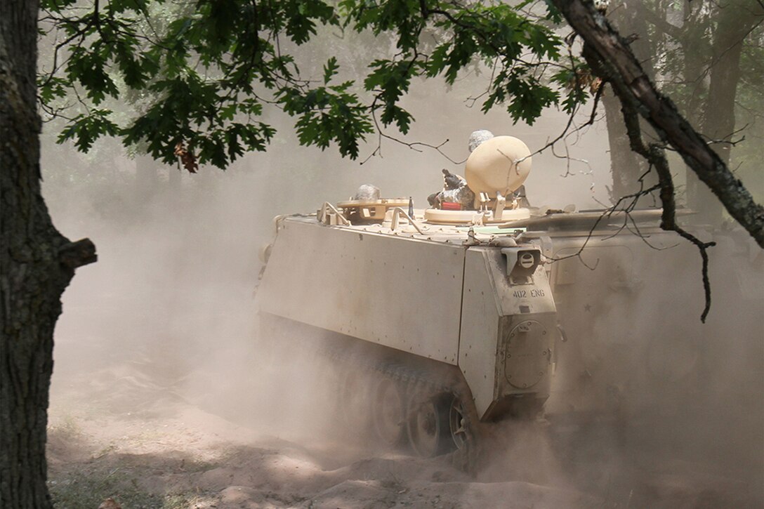 A moving combat vehicle kicks up dust clouds on a dirt road.