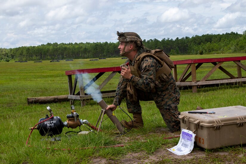 A service member kneels on the ground grasping a smoking mortar.