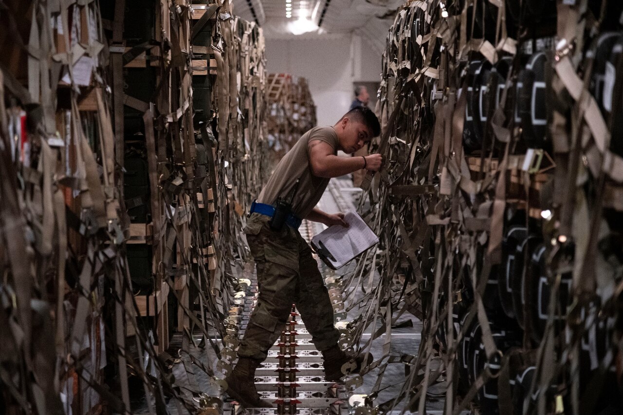 A service member tightens a strap to secure pallets of supplies inside an aircraft.
