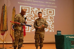 Male Soldier in green uniform speaks into a microphone while holding a baseball bat that is being presented to the male Soldier in green uniform standing next to him.