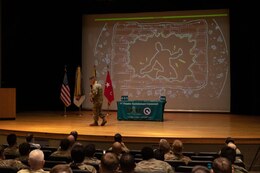 Male Soldier green uniform holds speaks into microphone and walks the stage as male and female Soldiers in green uniforms sit in seats in the audience and observe.