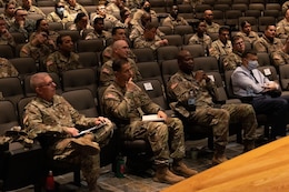 Male and female Soldiers in green uniforms sit in auditorium seats and look at the speaker on stage.