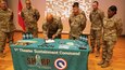 Male Soldier in green uniform uses pen to sign paper while male and female Soldiers in green uniforms stand around him and observe.