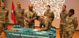 Male Soldier in green uniform uses a pen to sign a piece of paper as male and female soldiers in green uniforms stand around him and and observe.