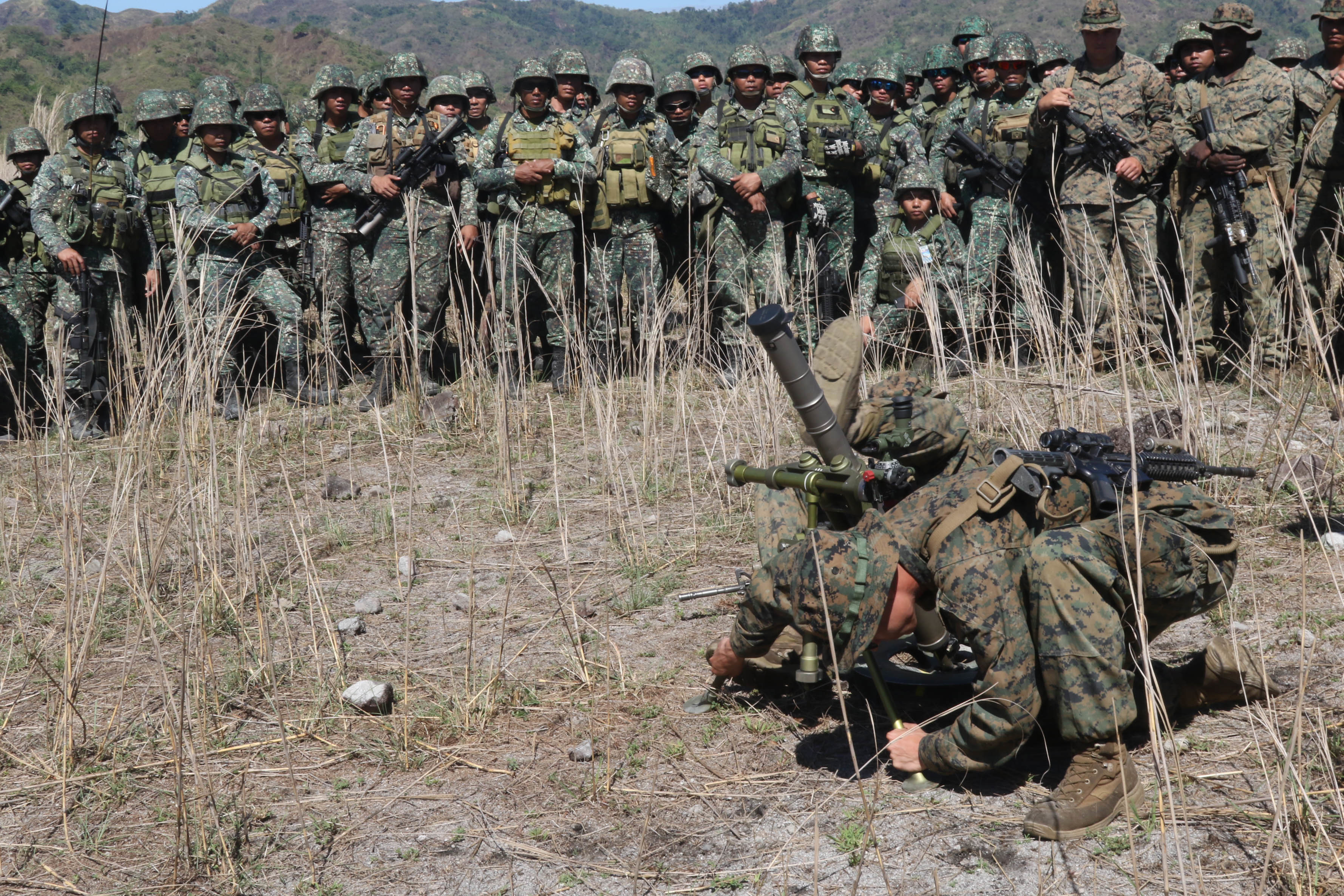 A line of Marines standing behind and looking at a Marine crouching on the ground.