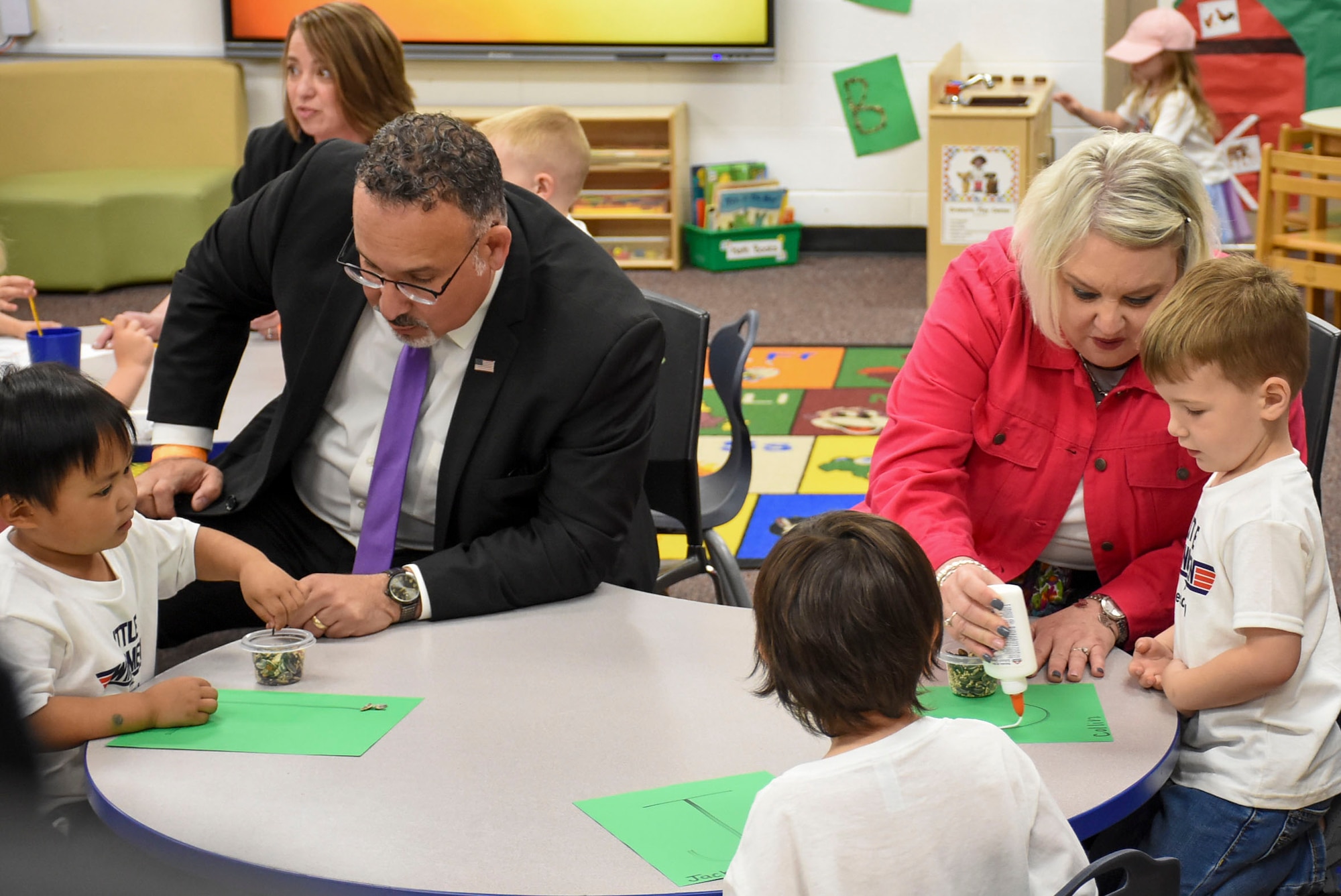 Dr. Miguel Cardona, wearing a purple tie to show support for military children, sits at a table with a woman in a red jacket and three preschoolers wearing white "Little Wingmen" t-shirts.