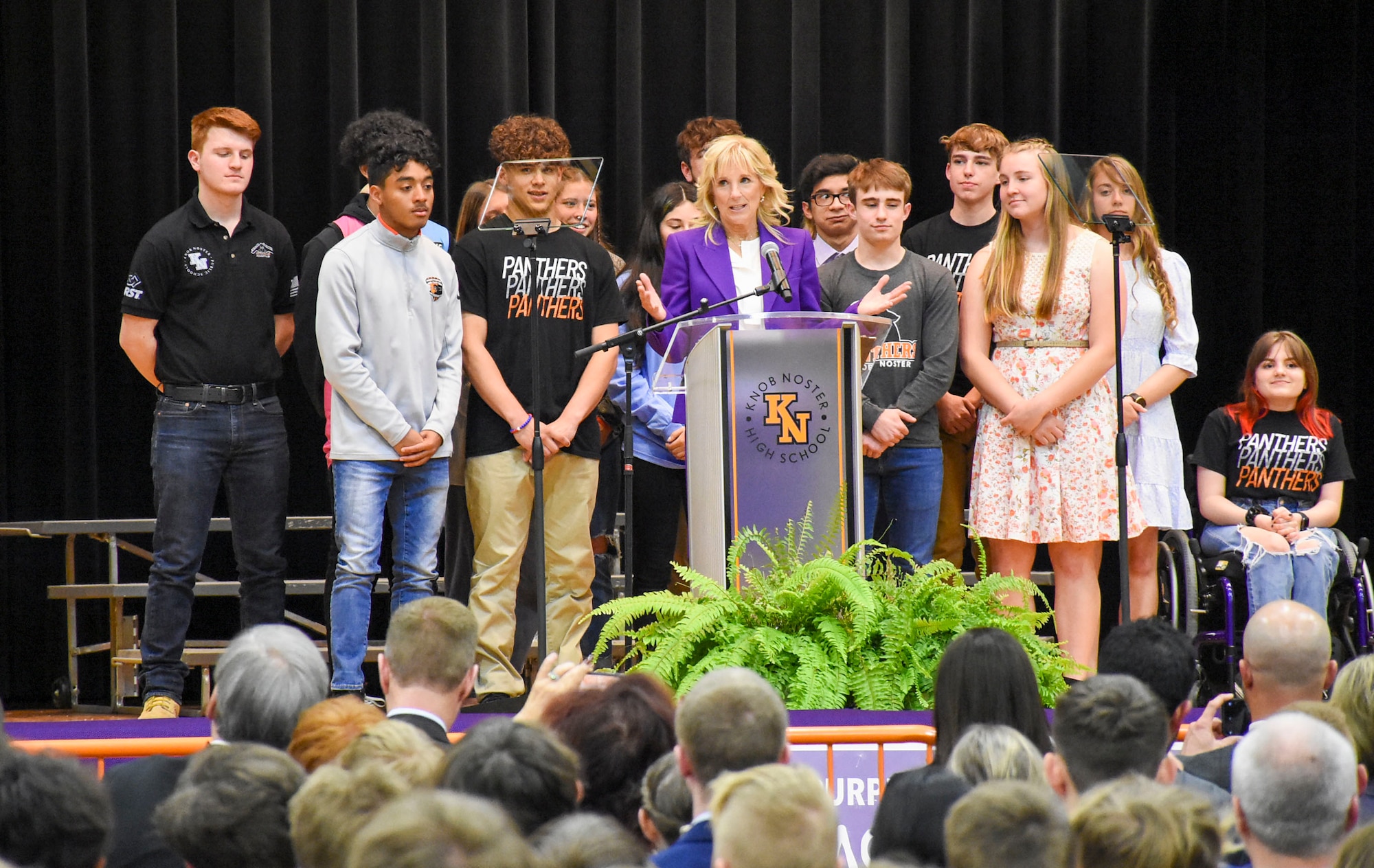 Dr. Jill Biden, wearing a purple blazer to show support for military children, speaks at a podium before a seated audience, while a group of high school students stands behind her.