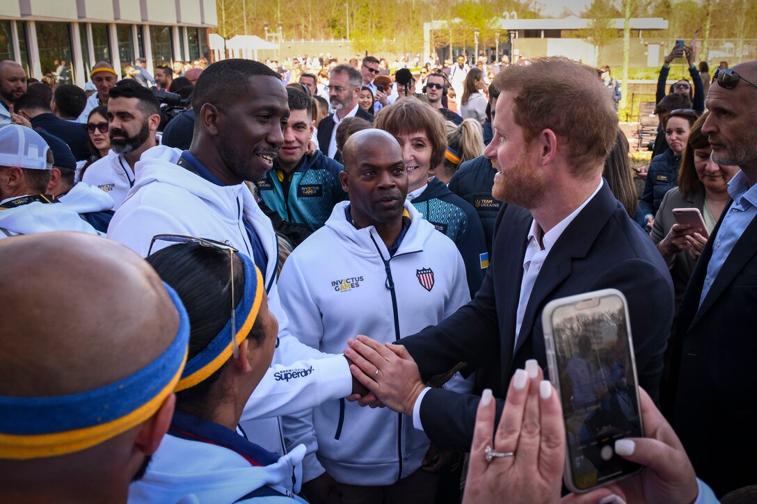 Prince Harry shakes hands with an airman while surrounded by a crowd.