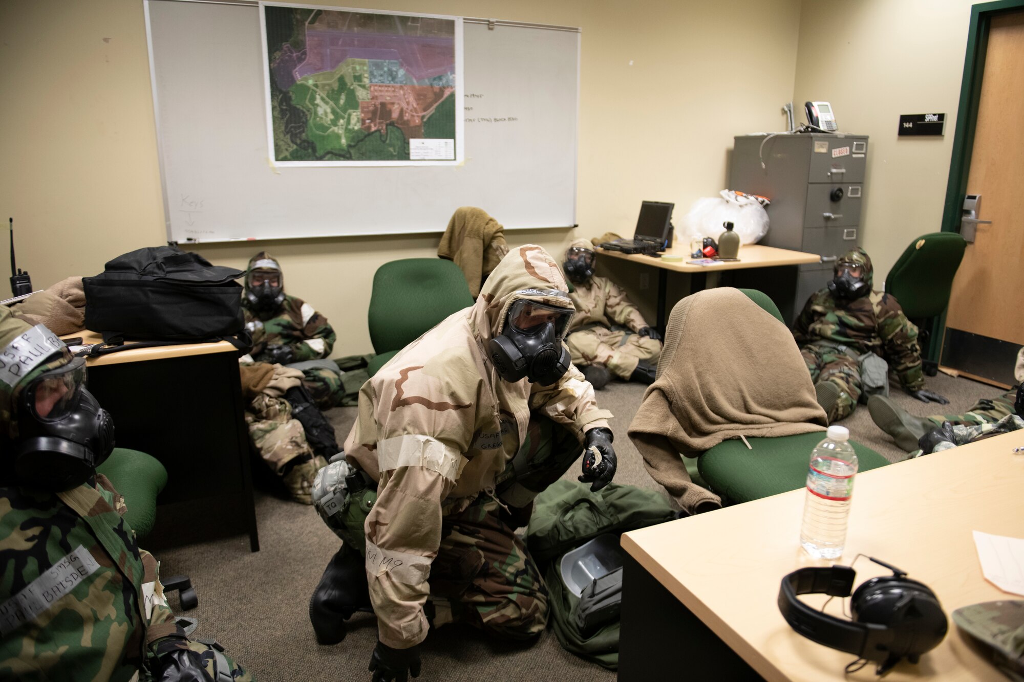 U.S. Airmen from the 60th Maintenance Squadron out of Travis Air Force Base, California, huddle for safety during a simulated chemical attack at the Alpena Combat Readiness Training Center, Alpena, Michigan, April 11, 2022. There are five Airmen pictured, siting or taking a knee on the floor of an office-like room.