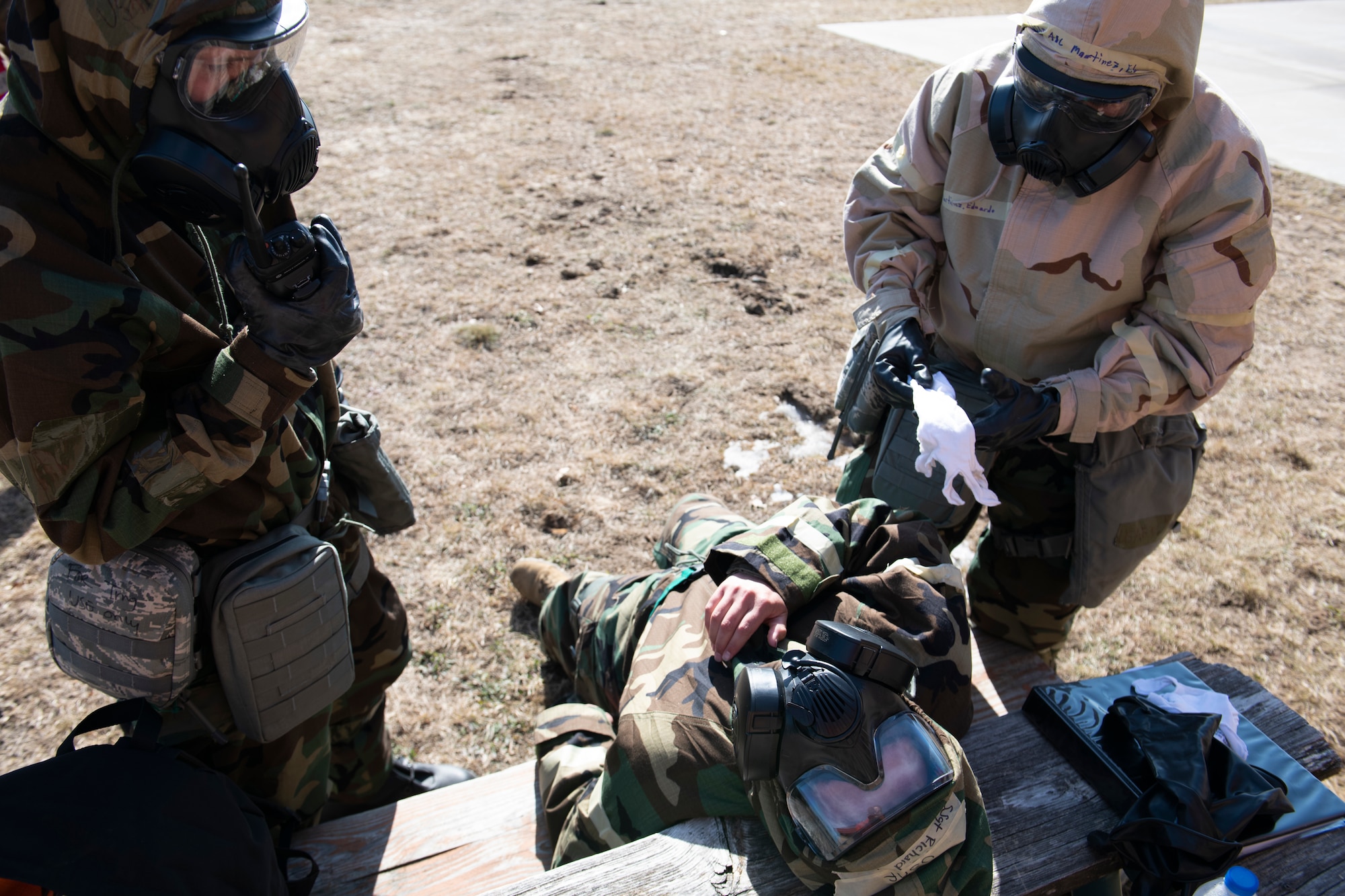 U.S. Air Force Staff Sgt. Jordan Harris, left, 60th Maintenance Squadron aero repair team leader, and Airman 1st Class Eduardo Martinez, 60th MXS aero repair team member, assess the condition of a 60th MXS Airman with simulated chemical burns at the Alpena Combat Readiness Training Center, Alpena, Michigan, April 12, 2022. The simulated burn victim is laidback on a picnic table as the other two Airmen are standing on both sides of him.