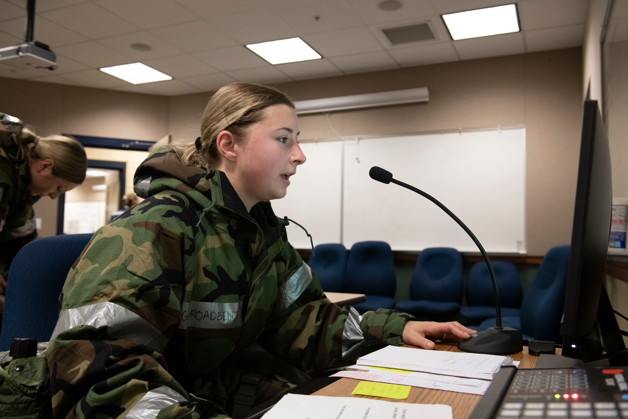 U.S. Air Force Staff Sgt. Charlotte Broadbent, 60th Air Mobility Wing Command Post controller, speaks into the giant voice system at the Alpena Combat Readiness Training Center, Alpena, Michigan, April 12, 2022. Broadbent is wearing MOPP gear, seated, and speaking into a small microphone.