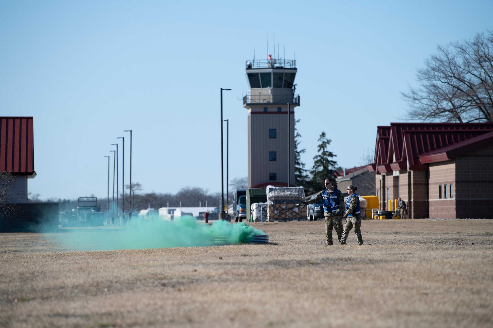 U.S. Air Force Tech. Sgt. Sean Minnis, left, 60th Air Mobility Wing Inspector General team member, throws a green smoke grenade to simulate a chemical attack as fellow IG teammate Staff Sgt. Kristen Patlan observes at the Alpena Combat Readiness Training Center, Alpena, Michigan, April 12, 2022. The Airmen are in a wide open grassy field with a hardened dugout which the smoke grenade is thrown in. In the distance is an air traffic control tower.
