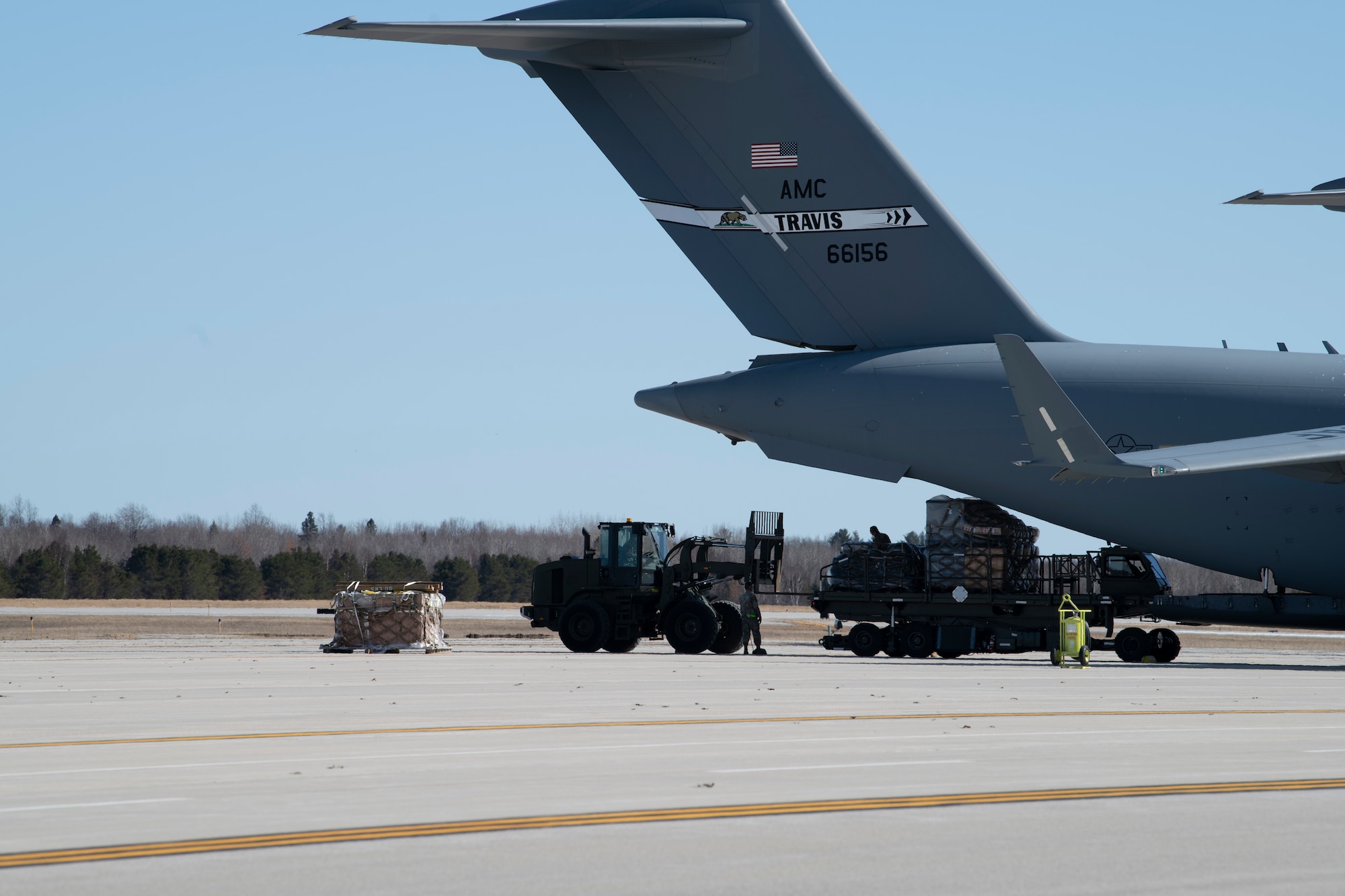 U.S. Airmen from the 60th Aerial Port Squadron out of Travis Air Force Base, California, off-load cargo from a C-17 Globemaster III at the Alpena Combat Readiness Training Center, Alpena, Michigan, April 10, 2022. A fork-lift and k-loader are pulling cargo out of the back of C-17.