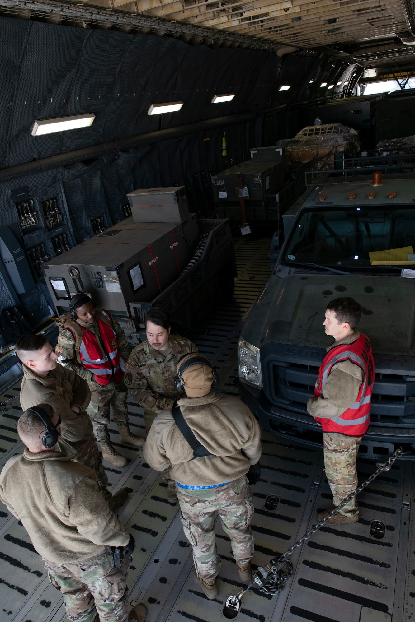 U.S. Airmen from the 60th Aerial Port Squadron out of Travis Air Force Base, California, discuss off-loading plans in a C-5M Super Galaxy cargo compartment in preparation for Exercise Roundel Perun 22-01 at the Alpena Combat Readiness Training Center, Alpena, Michigan, April 10, 2022. A group of six Airmen are standing in the front portion of the cargo compartment and compartment is filled with equipment.