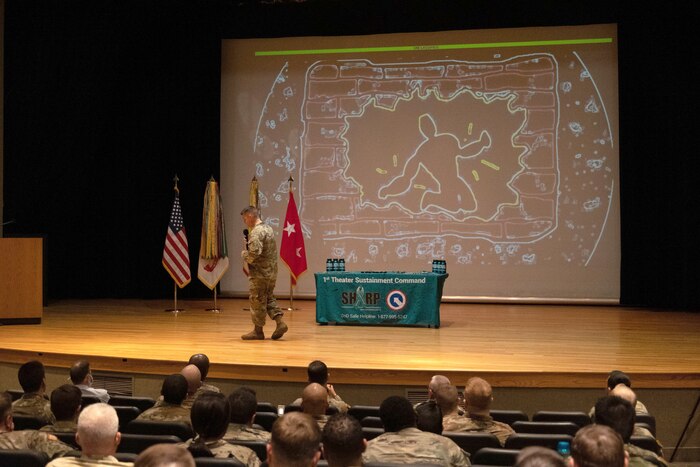 A soldier walks across a stage while an audience watches.