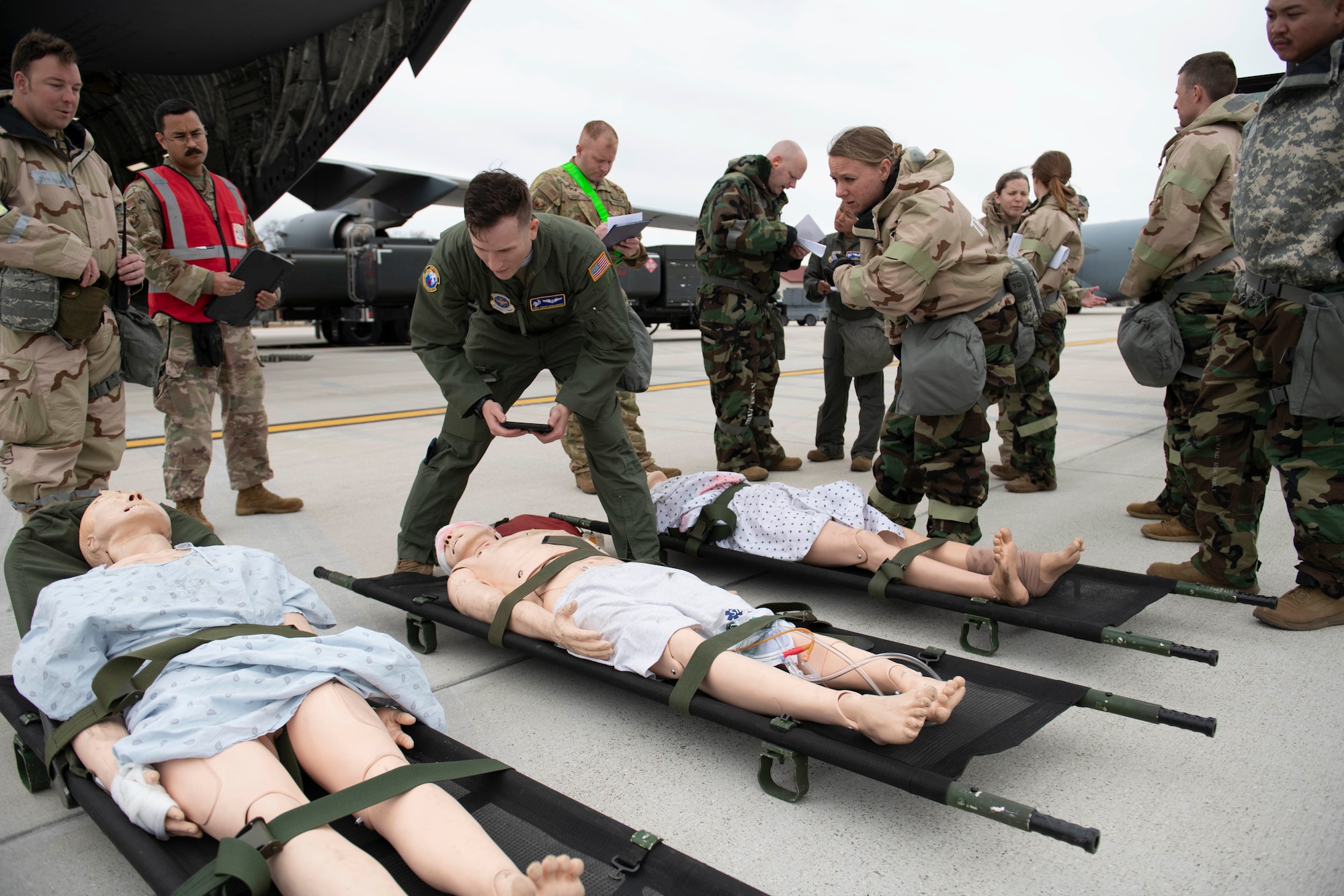 U.S. Air Force Tech. Sgt. Coley Kicklighter, third from left, 60th Aeromedical Evacuation Squadron AE technician, verifies the medical information of simulated patients before they load into a C-17 Globemaster III from Travis Air Force Base, California, at the Alpena Combat Readiness Training Center, Alpena, Michigan, April 10, 2022. There are three simulated patients on the ground and they're surround by Airmen dressed in MOPP gear.