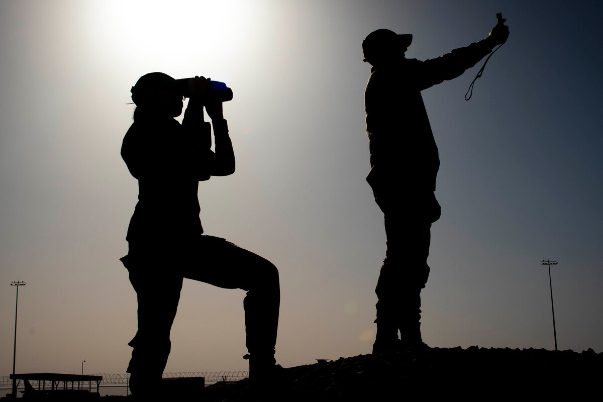 Two airmen shown in silhouette while one uses binoculars and the other uses a weather instrument.