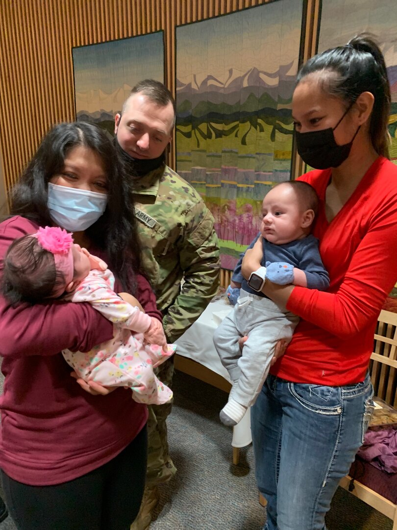 Women in forefront holding infants with a soldier looking on in the background. Mountain quilts hanging on wall.