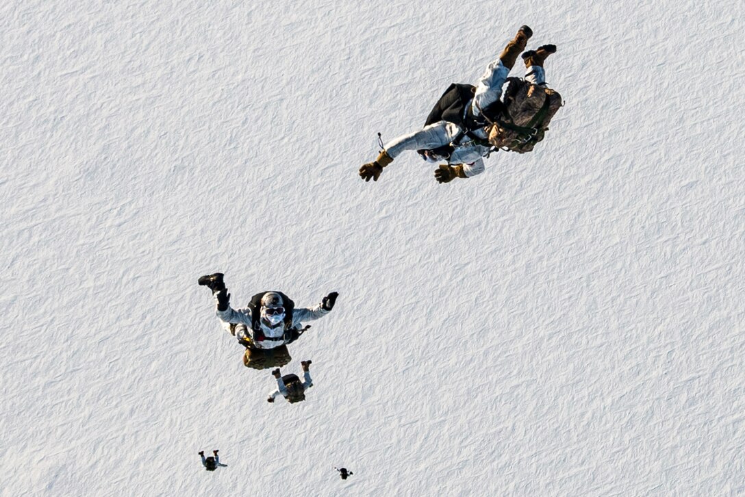Four sailors free fall with parachutes over snowy surface.
