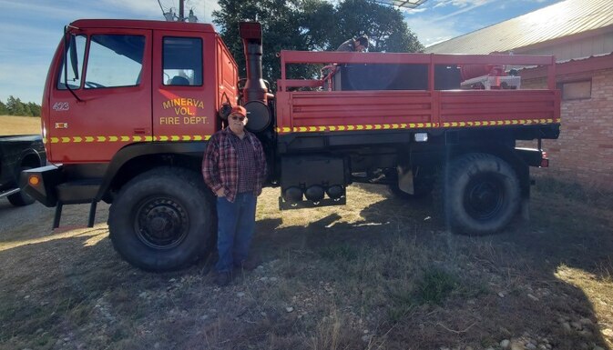 Fire Chief Ernie Glenn of the Minerva Volunteer Fire Department in Texas stands next to a former military truck. The truck has been converted into a fire truck and painted red.