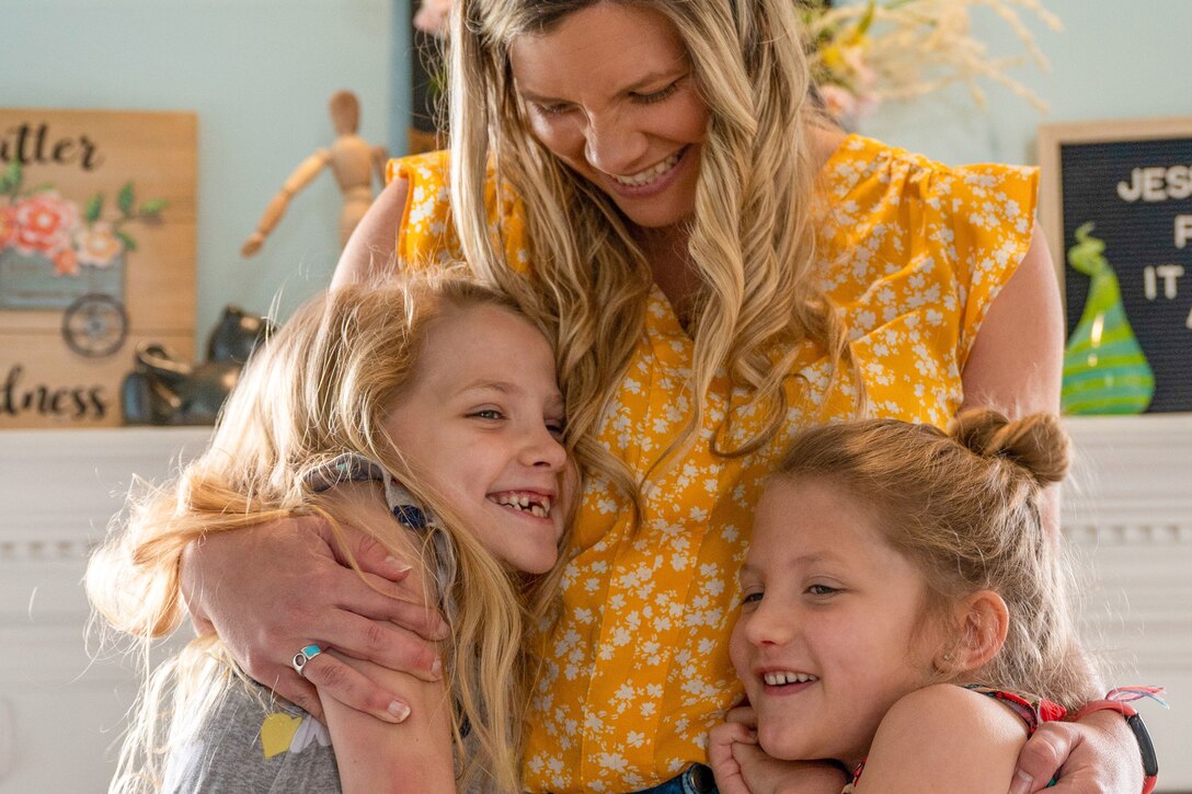 A smiling woman puts one arm each around two smiling girls.