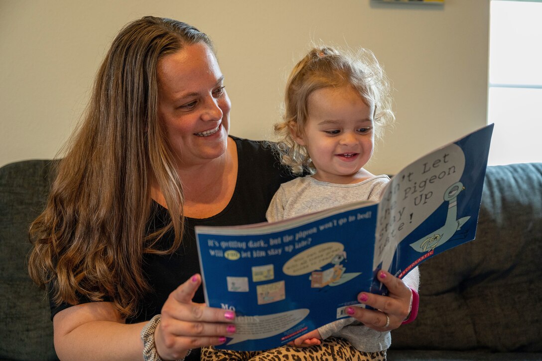 A mother reads a book to a toddler sitting on her lap on a couch.