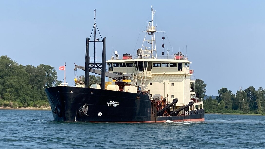 A large ship, called a hopper dredge, travels along a river on a sunny day