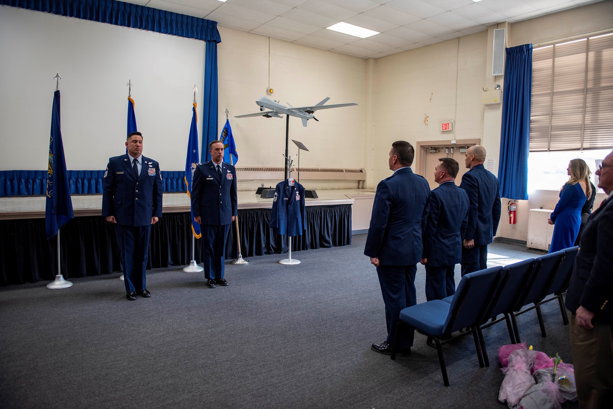 Two men in Air Force enlisted dress blues stand at attention in front off a military audience in an auditorium.