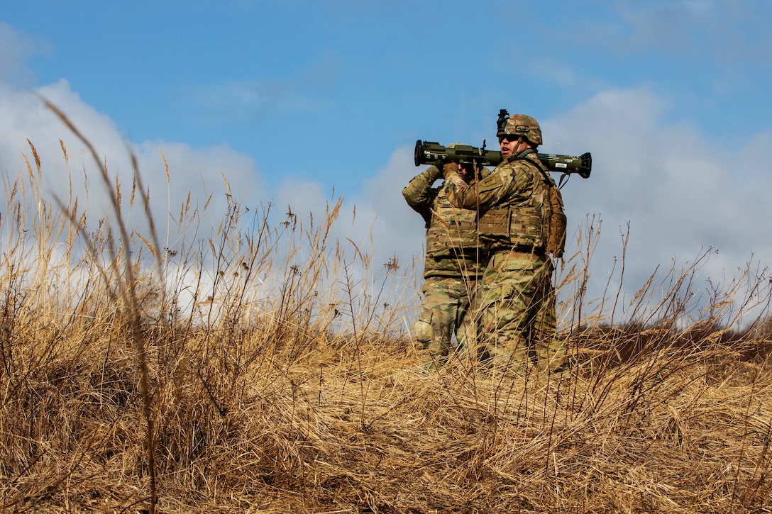 One soldier holds an anti-tank shoulder weapon while another soldier adjusts it.