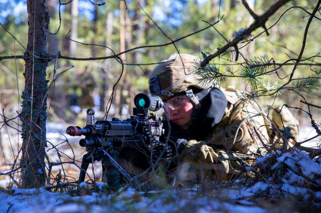 A soldier lying on the ground looks through the scope of an automatic rifle.