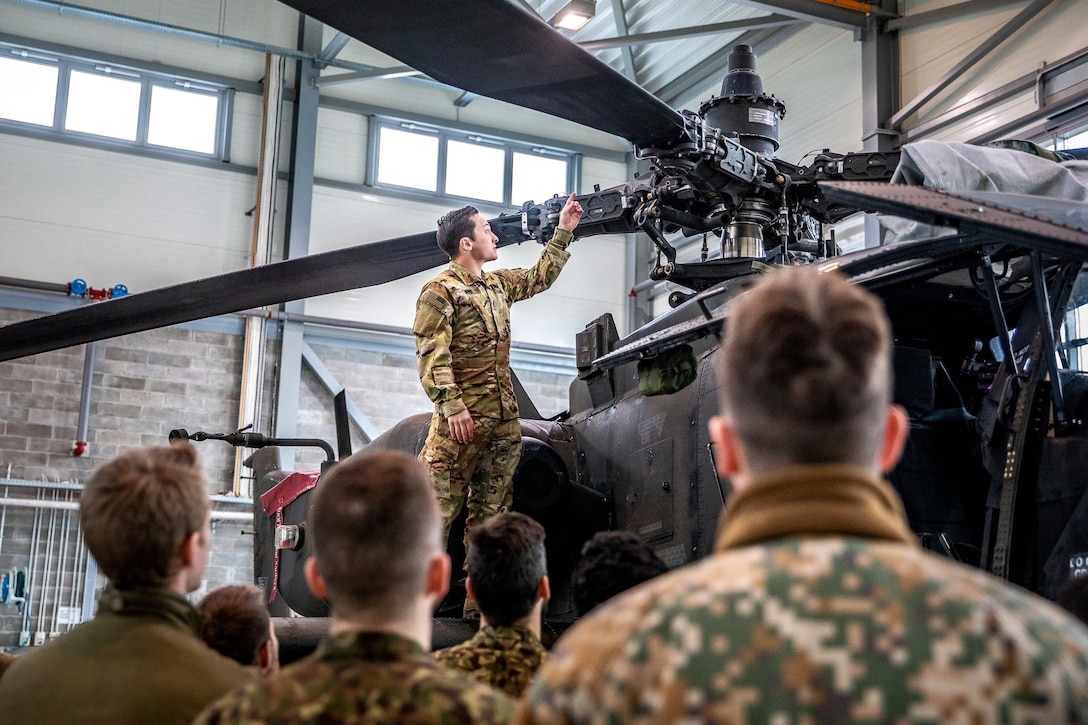 A soldier stands on the wing of a helicopter while gesturing to its rotor in front of a crowd.