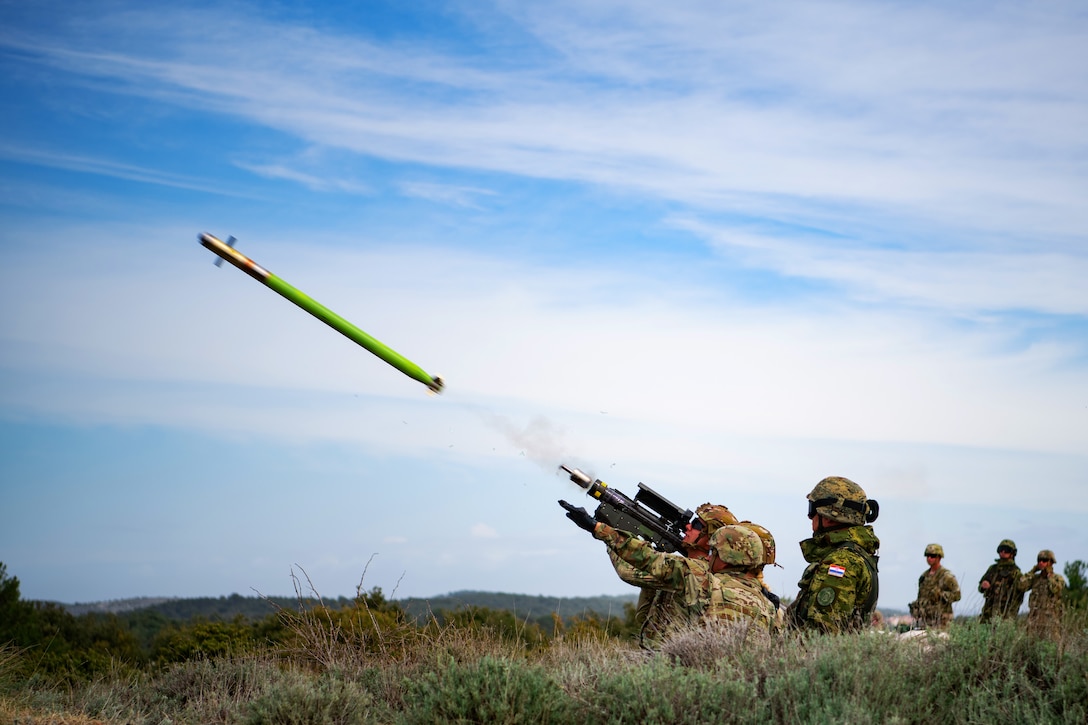 Soldiers in a kneeling position fire a shoulder-mounted missile weapon while three other soldiers observe in the background.