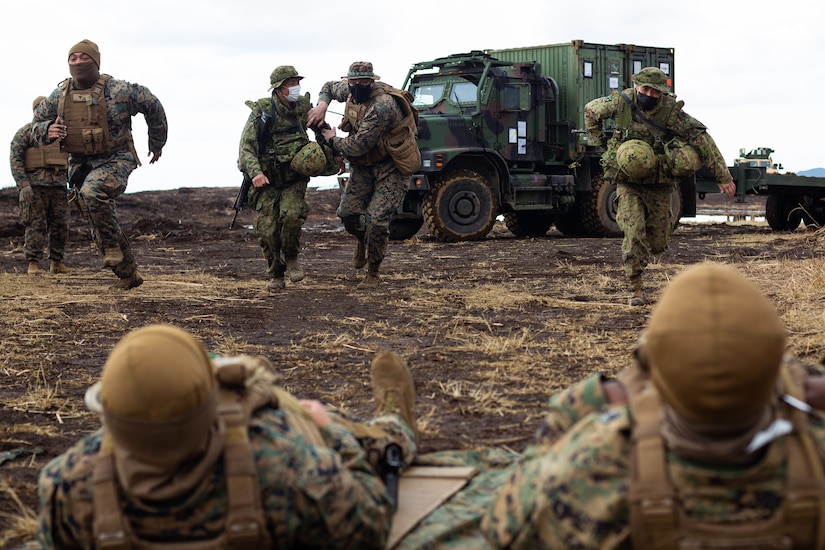 Medical personnel run toward two service members lying on the ground.