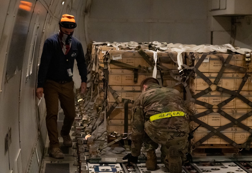 Two individuals, one in a military uniform, affix straps to cargo palettes in the rear of an aircraft.