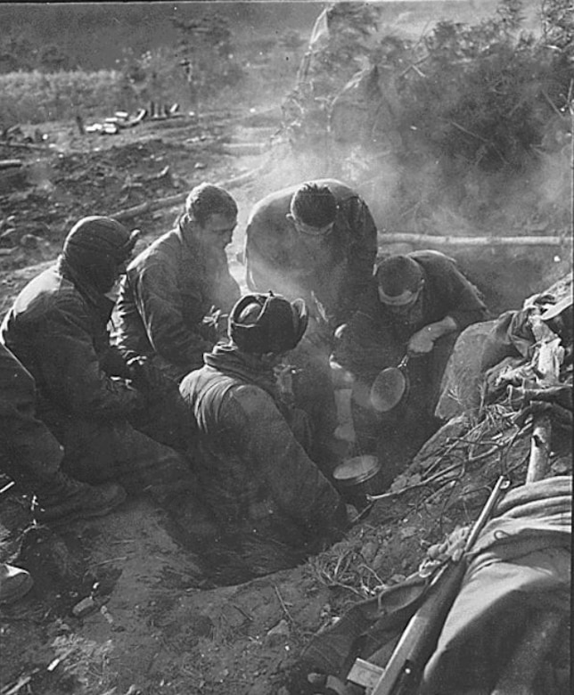 Five men look toward a pot cooking something in a foxhole on a battlefield.