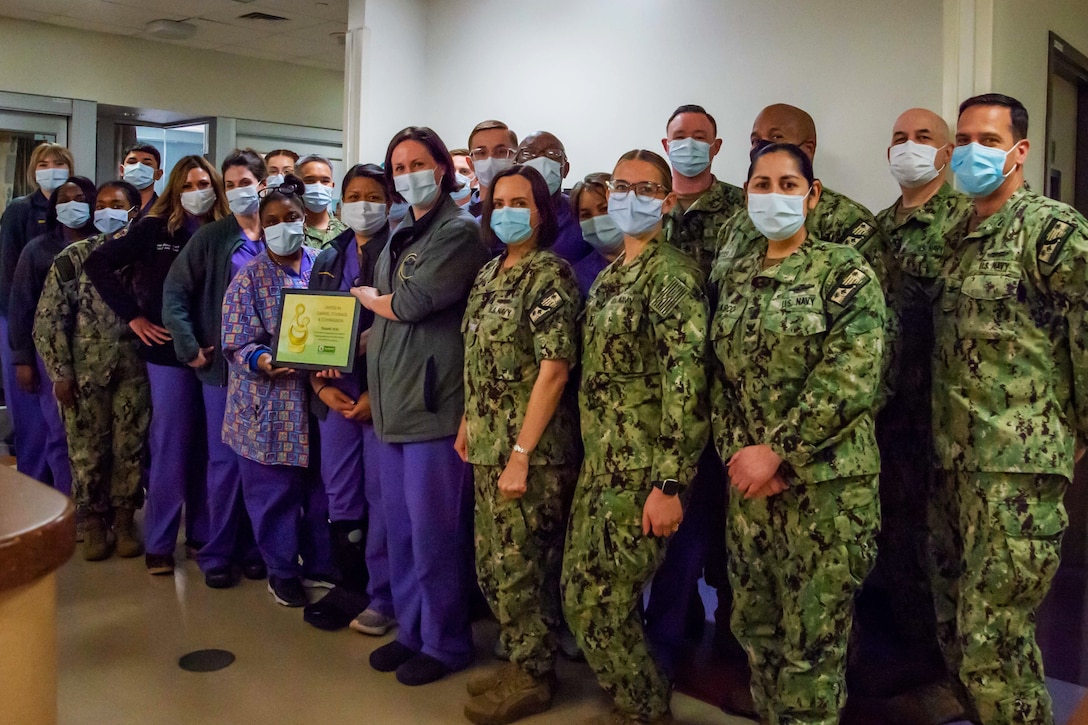 A large group of medical staff and Navy sailors display an award while posing for a group photo inside a hospital.