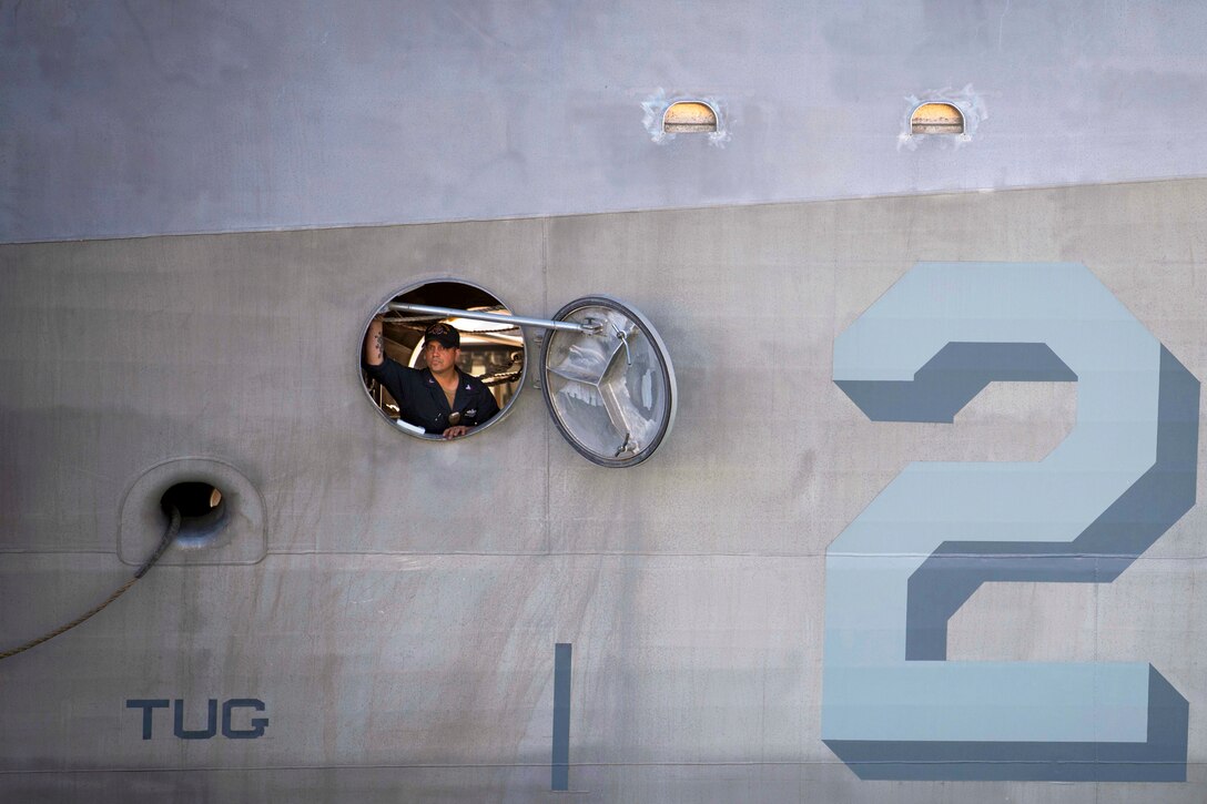 A sailor peeks out of a circular opening on the hull of a ship.