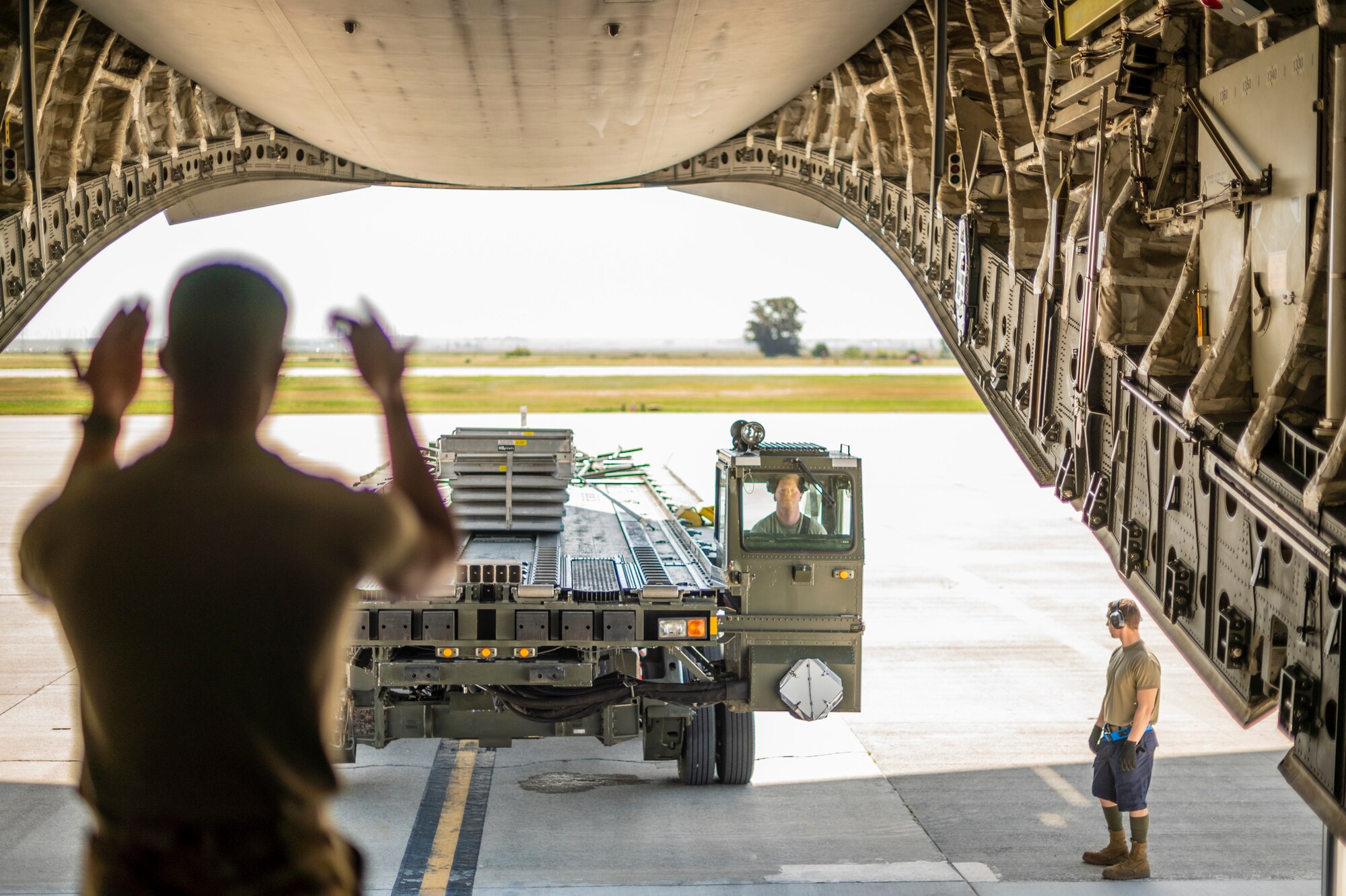 A man gestures to a large military vehicle that is used to load pallets on to large military aircraft, in side of a C-17 Globemaster III, a large Air Force aircraft.