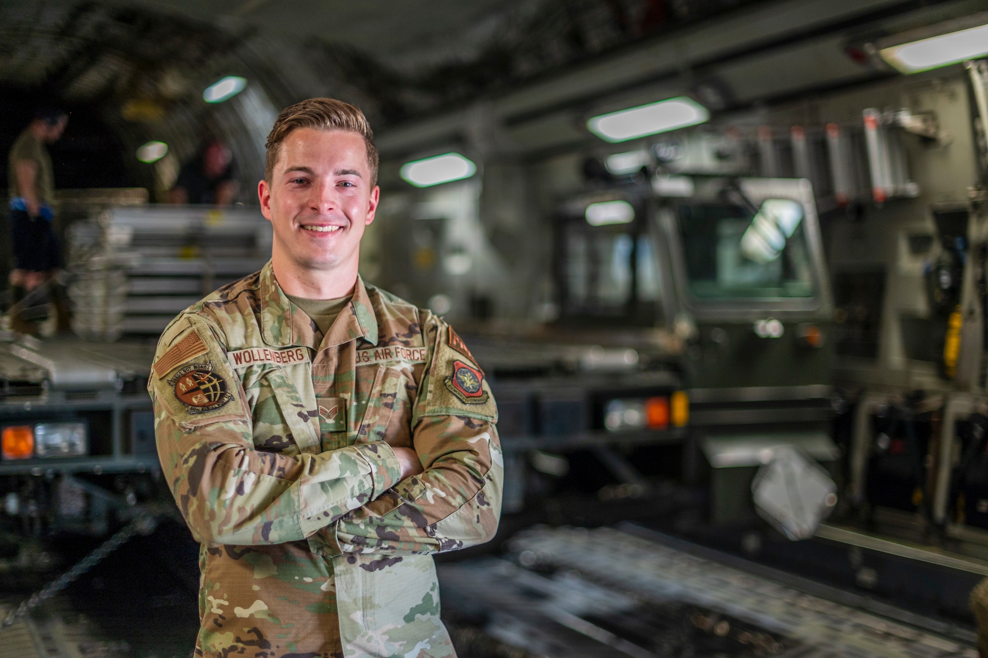 A man poses for a picture in front of a large military vehicle that is used to load pallets on to large military aircraft, in side of a C-17 Globemaster III, a large Air Force aircraft.