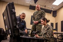 Two uniformed marines and one civilian gather around an open box while one inspects a camera.