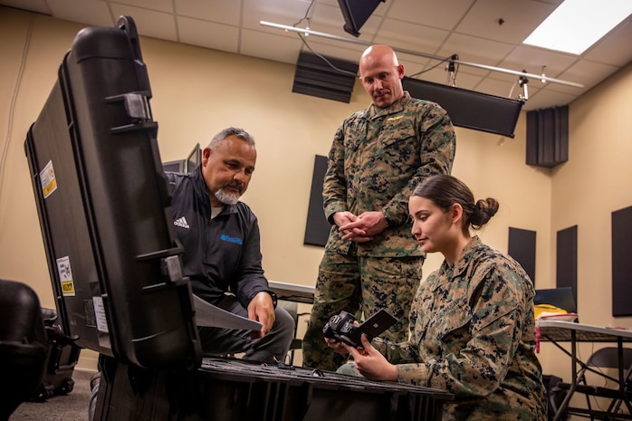 Two uniformed marines and one civilian gather around an open box while one inspects a camera.