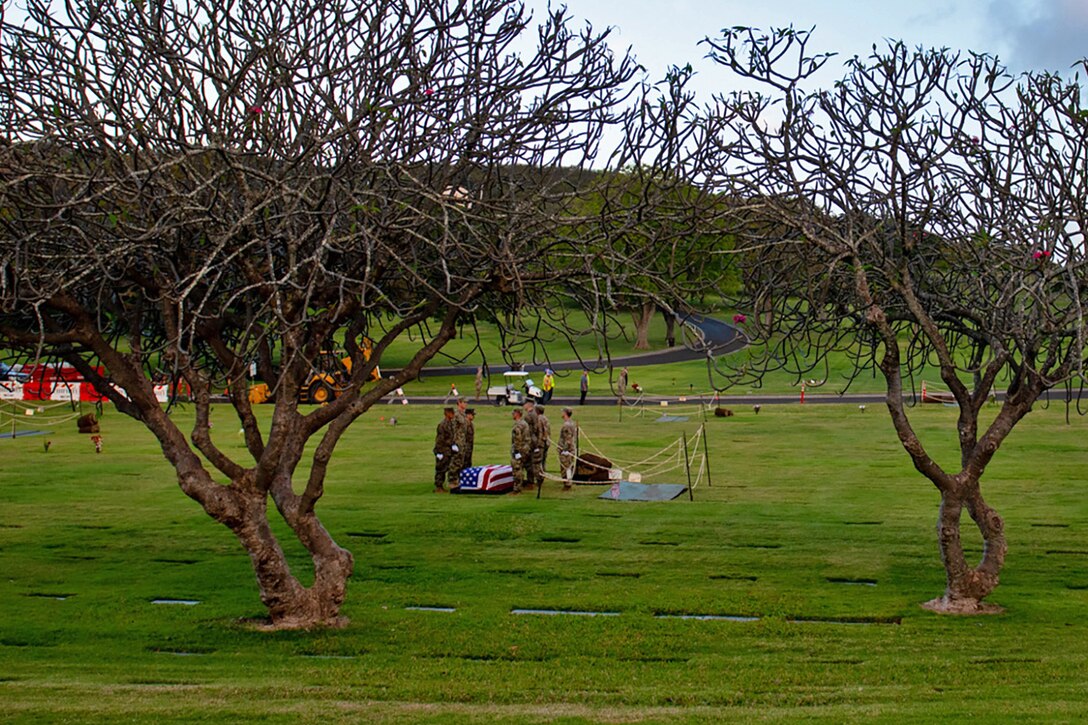 A group of service members framed by two trees stand by a grave in a cemetery.