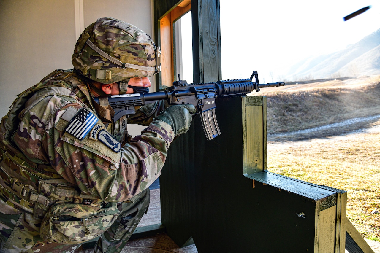 A soldier, kneeling behind cover, shoots a rifle.