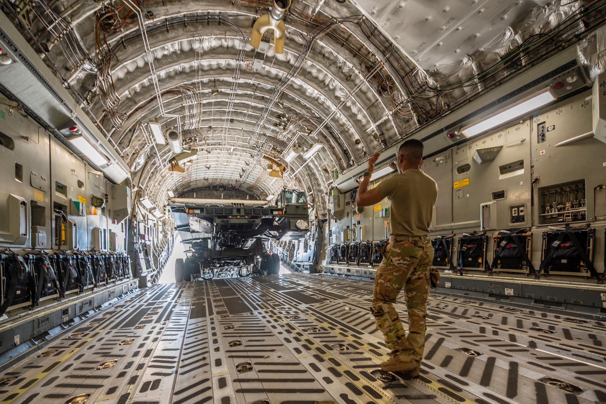 A man gestures towards a large military vehicle that is used to load pallets on to large military aircraft, in side of a C-17 Globemaster III, a large Air Force aircraft.