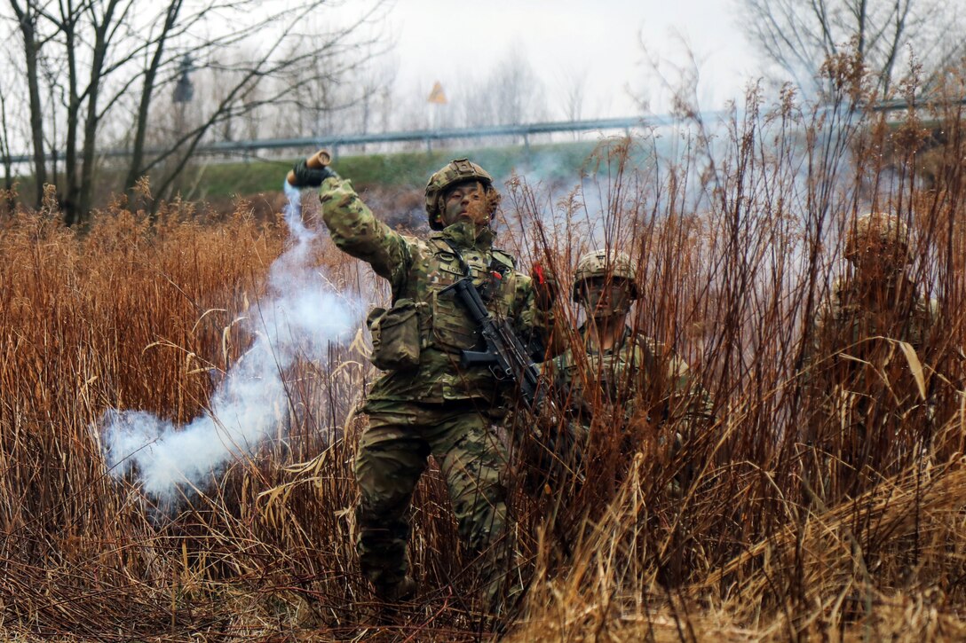 A paratrooper throws a smoke grenade while two other solders stand behind them.