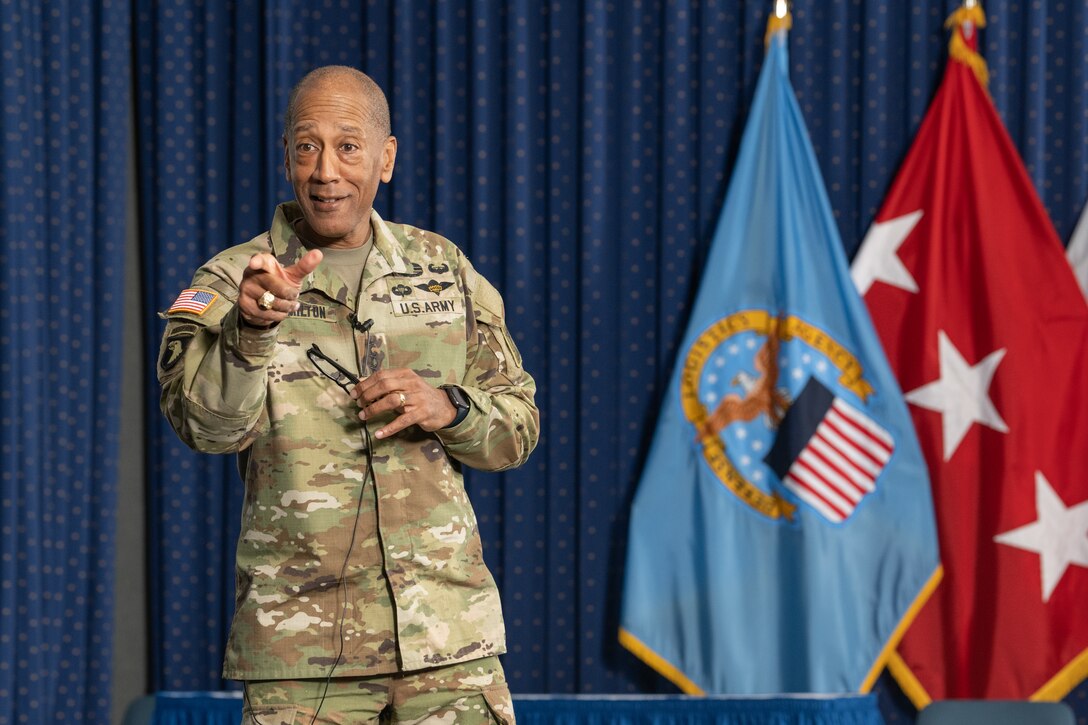 Black man in camo uniform stands in front of flags.