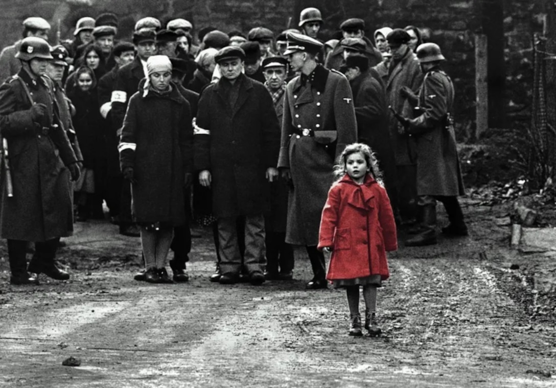 Nazi soldiers escorting a group of Jewish people on a street with a little girl in a red coat in the front. The whole scene is black and white except for the little girl's red coat.