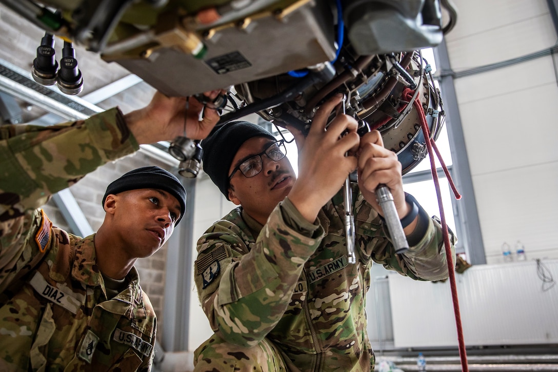 Two soldiers work on an engine of an AH-64D Apache Longbow helicopter.