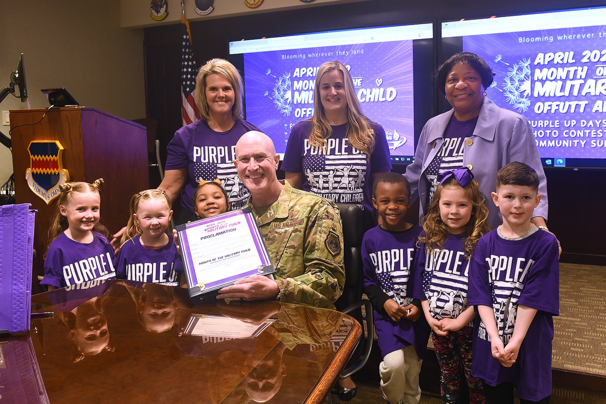 A group of children and adults stands around a man in uniform sitting down and holding up a proclamation.